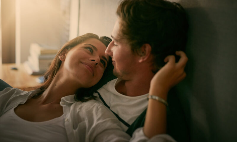 frau und mann liegen gemeinsam im bett und frau schmiegt sich liebevoll an ihn und schaut ihm in die augen